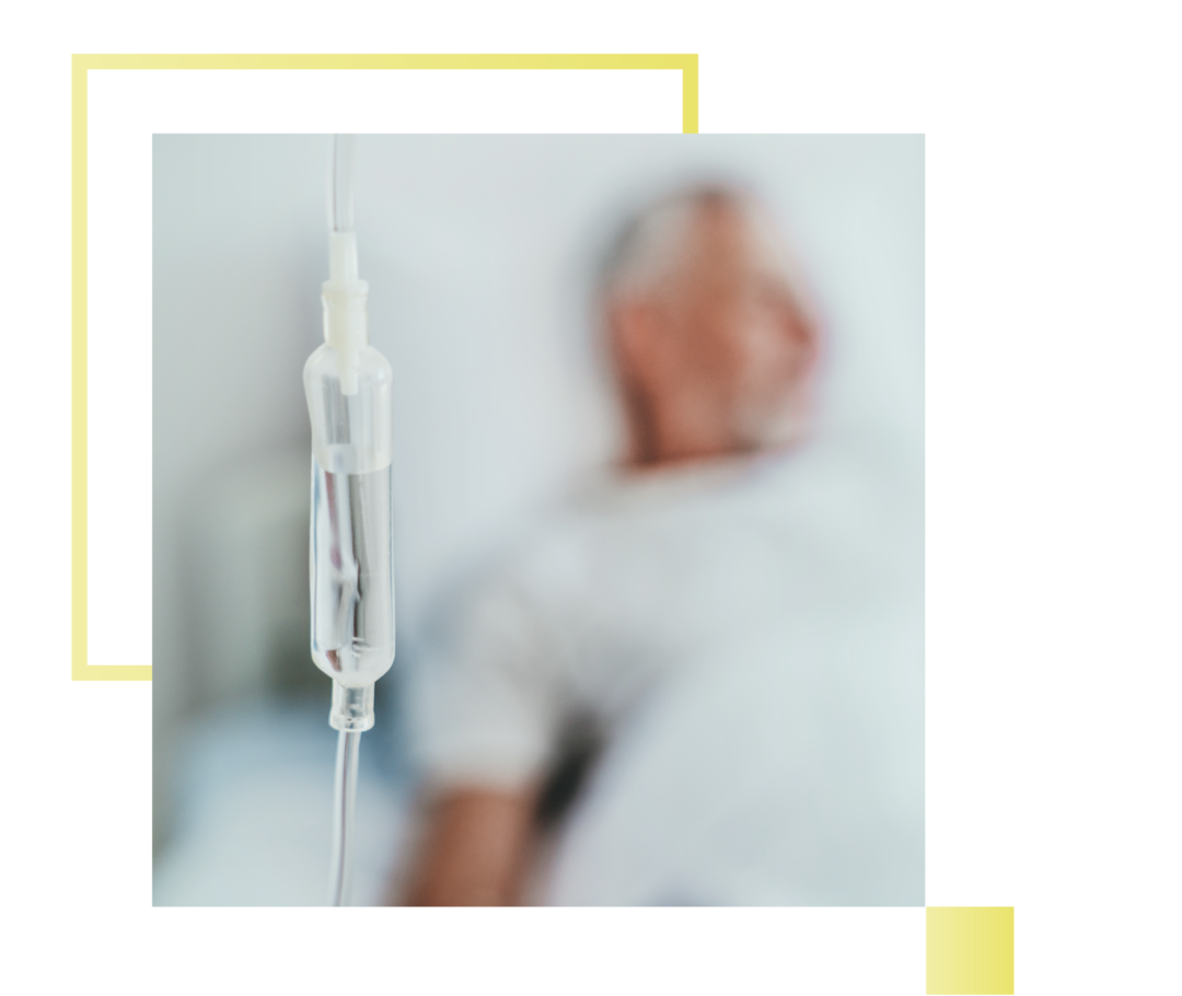 Abgebildet ist eine Infusionsflasche. Im Hintergrund ist ein Patient zu sehen.