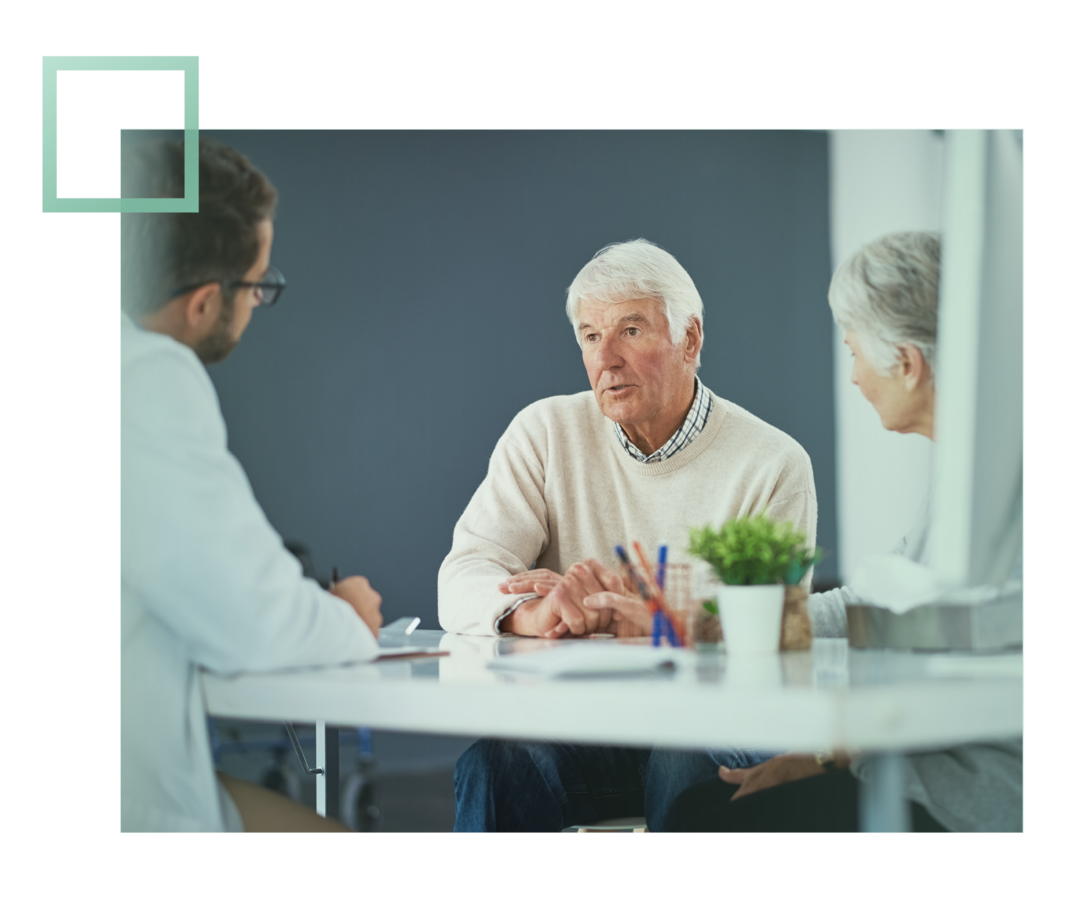 Leben mit fortgeschrittenem Prostatakarzinom: Ein älterer Herr und seine Frau sitzen mit einem Arzt an einem Tisch. Der Mann unterhält sich mit dem Arzt. Ein älterer Herr und seine Frau sitzen mit einem Arzt an einem Tisch. Der Mann unterhält sich mit dem Arzt.